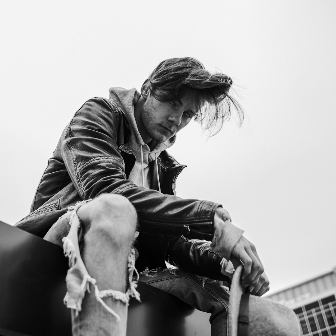 Black and white photography of a man with a leather jacket and ripped jeans sitting on top of a building looking down in the style of minimalism and portrait.