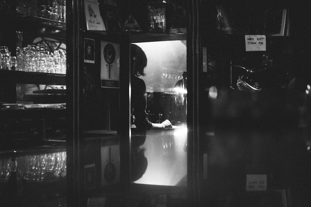 In the dark corner of an empty bar, there is only one woman sitting alone at her table in front of me. The mirror on top shows my reflection as I look into it and see another person’s face. A glass cabinet with various glasses stands next to him. In Japanese photography style, the foreground is blurry and the background is clear. Black and white tones create contrast between light and shadow. Using Leica camera shooting technology for film and television works. It has strong emotional impact.,,in –ar 128:85