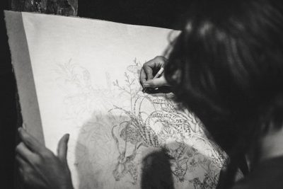 A black and white photo shows an artist drawing on paper with a pencil in hand, drawing patterns on fabric with a side view in a dark room. It is a close up, low angle shot focused on the cloth with intricate patterns while the blurred background shows other artists working around her in the style of the first artist. --ar 128:85