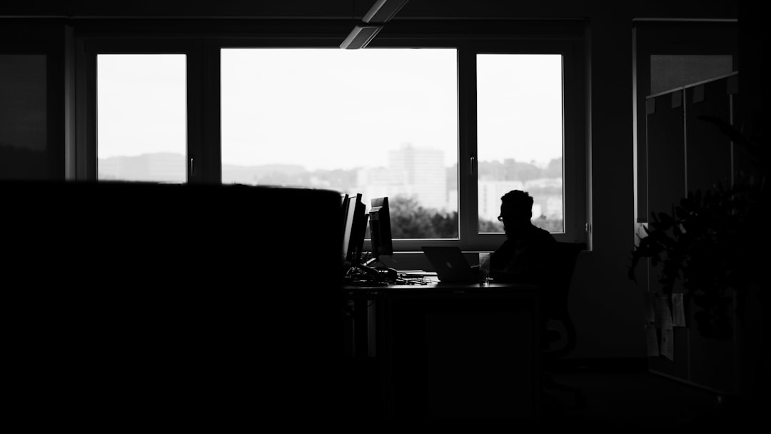 Silhouette of a person sitting at a desk in a dark office with a window showing a cityscape. Black and white photo. Silhouettes of people working on computers. Monochrome photography. Minimalist interior design of a modern home or business space. High contrast, natural lighting, Nikon D850 camera with an f/2 lens, capturing the intricate details of the silhouette against the stark background in the style of the photographer. –ar 16:9