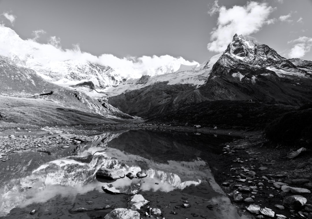 Black and white photography of the view from little lake in mountain valley with reflection, snowcapped mountains, rocky shore, clear sky, mountain peak, shadow, high contrast, rocks, water ripples, alpine landscape, –ar 64:45