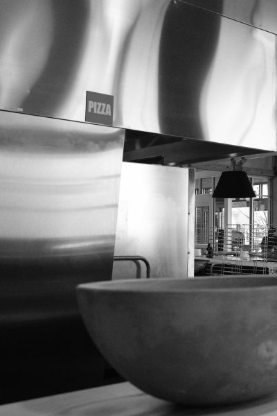 Black and white photograph of an empty pizza oven, blurred kitchen in the background with "PIOPENA PCHANTI", and a large bowl on one side. The focus is on capturing the sleek design of stainless steel surfaces, emphasizing modernity within a commercial environment in the style of the artist. --ar 85:128