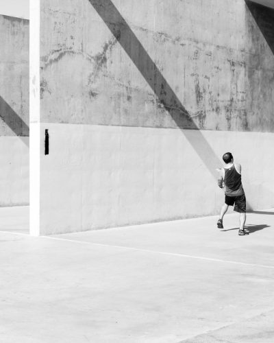 A black and white photo of an adult playing tennis in a parking lot against a concrete wall, with strong shadows and geometric shapes. It is a minimalist photography piece with a simple composition showing a full body shot from a low angle camera view, with sharp focus on the subject in natural sunlight. --ar 51:64