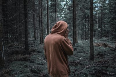 A man in a brown hoodie standing alone in the forest, with his back to the camera, with dark color grading in a hyper realistic style. --ar 128:85