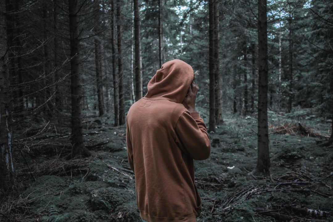 A man in a brown hoodie standing alone in the forest, with his back to the camera, with dark color grading in a hyper realistic style. –ar 128:85