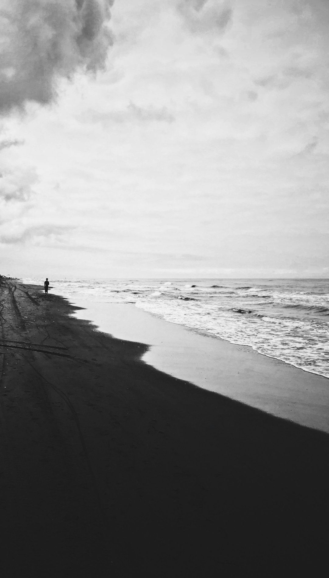 Black and white photography of the beach in perspective, with waves crashing onto sand. In front is an empty black beach with shadows on it, while behind there’s a person walking along its edge. The sky above is cloudy, adding to the contrast between lightness and darkness. This scene captures the serene beauty of nature in the style of focusing attention on both water and land, creating a peaceful atmosphere. Black background, white border. –ar 73:128