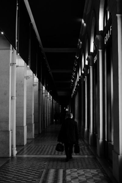 A man with his back turned carrying two bags walks along the corridor of an underground shopping center in Paris, illuminated by streetlights. The scene is captured from behind and has a black and white color scheme. It was taken at night using a Canon EOS camera with an f/20 aperture setting to capture details and create a contrast between lightness and darkness. --ar 85:128