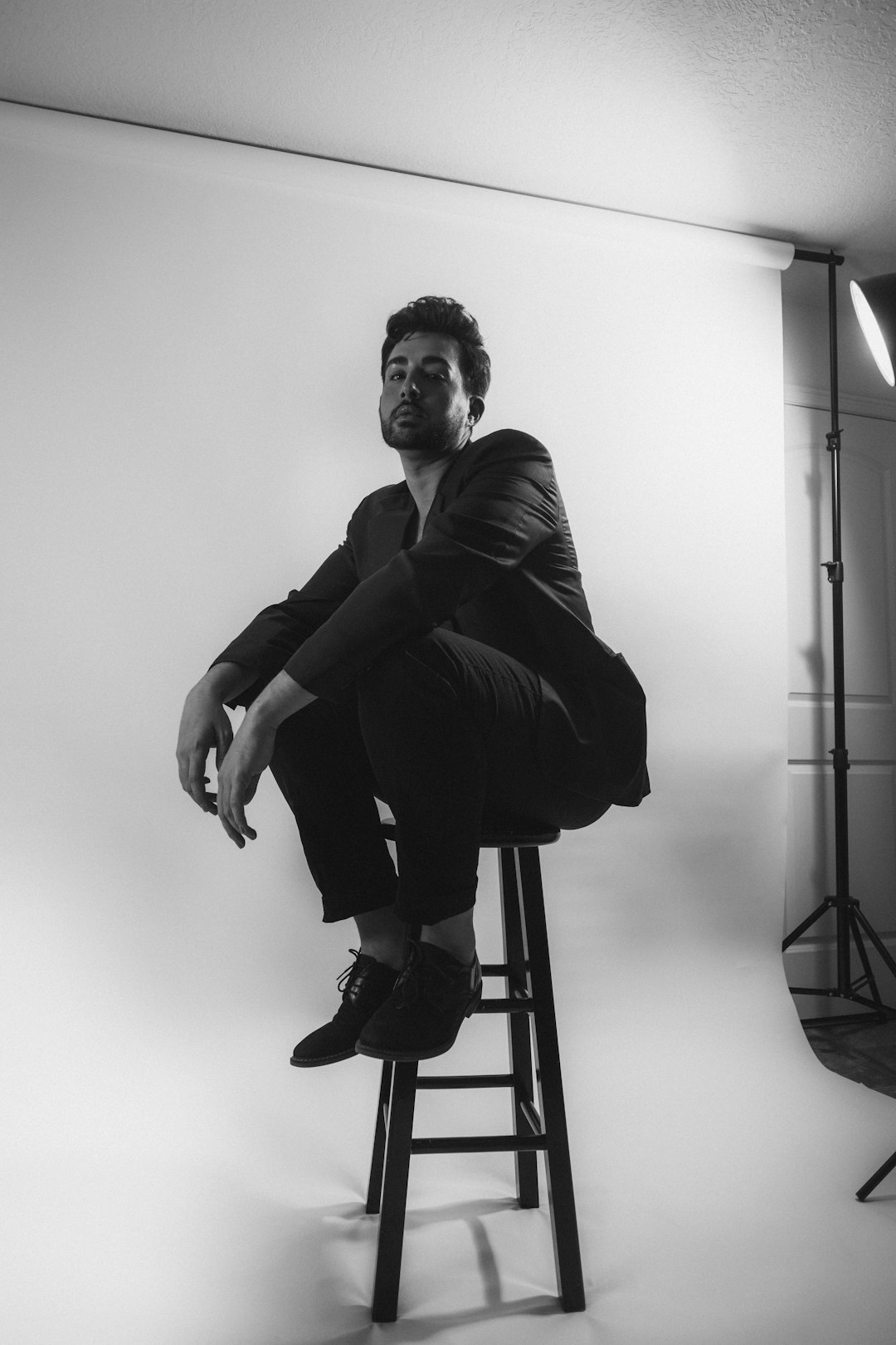 A black and white portrait of an actor sitting on a bar stool in a photo studio, with a white background, hard light, high contrast, a medium shot, in the style of fashion photography, in a raw style, wearing a suit jacket and pants, barefoot. –ar 85:128