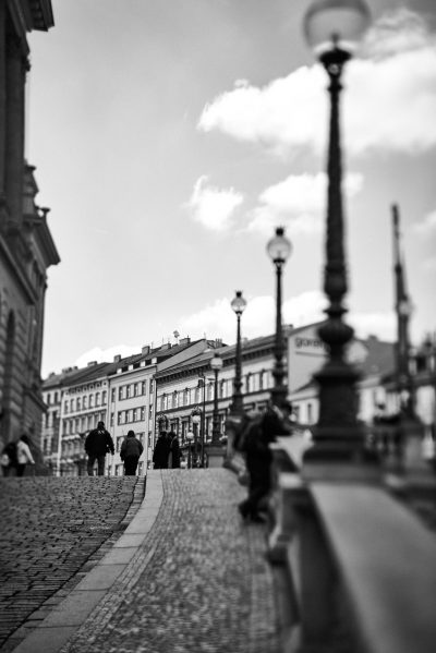 Black and white street photography of the city of Warsaw, Poland featuring street lamps, people walking on cobblestone streets with buildings in the background. The photos have a depth of field effect with a blurry foreground and are captured in a hyper realistic style reminiscent of a Leica M6 with a Summilux f/2 lens. Soft lighting and high resolution provide super detailed images. --ar 85:128