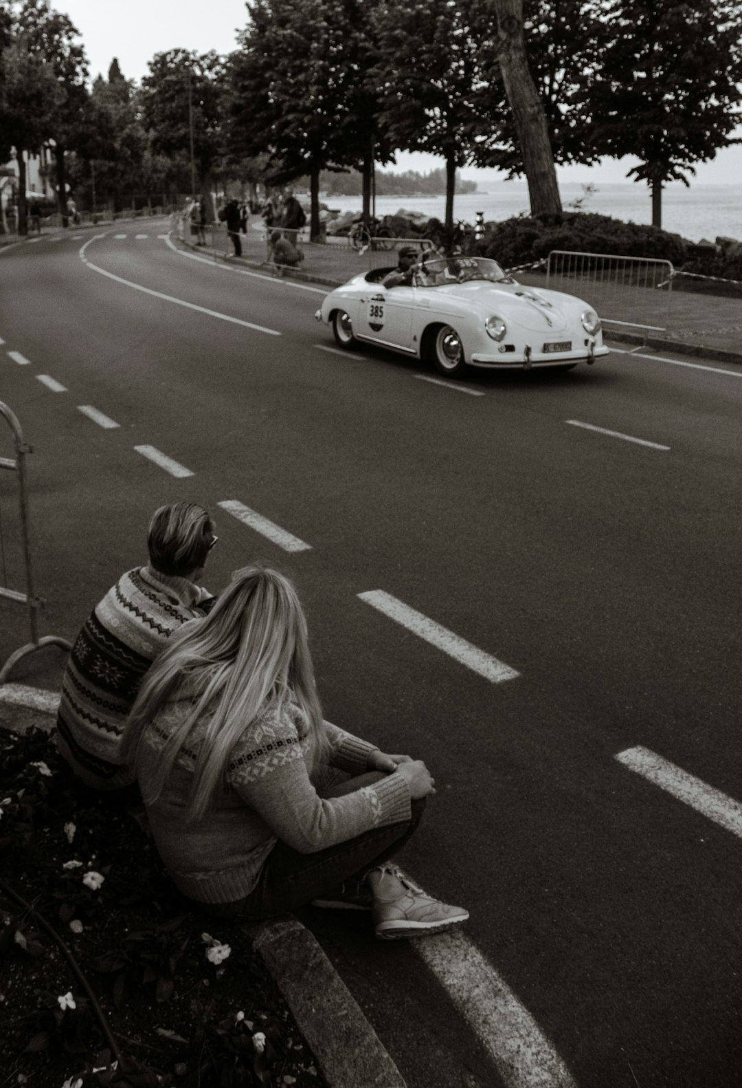 Black and white photography of a blond woman sitting on the sidelines in Lausanne with her boyfriend watching a vintage Porsche race car, with highly detailed style. –ar 87:128