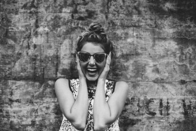 A black and white photograph of an excited woman wearing sunglasses standing in front of a textured wall, covering her ears with both hands, laughing heartily with her mouth wide open showing teeth, wearing a stylish summer dress with her hair tied back in a bun, having a vintage feel, with high contrast between the subject's skin color and the background texture, shot on a Leica M3 film camera in the style of street photography. --ar 128:85