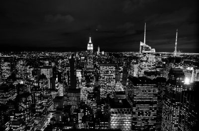 A black and white photograph of the New York City skyline at night, with skyscrapers illuminated by lights against a dark sky. The cityscape is bustling with activity, showcasing iconic buildings like Empire State Building and others in all their glory. Shot from an aerial perspective to capture its vastness and scale. High resolution photography in the style of highresolution photography. --ar 32:21