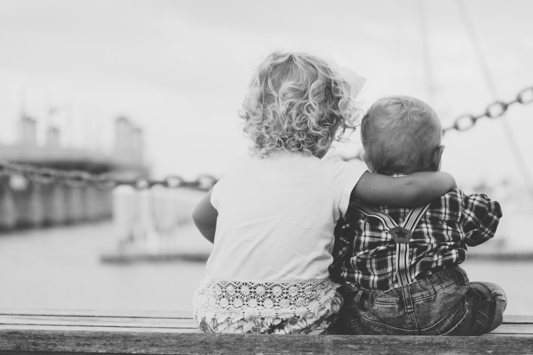 A black and white photo of two children sitting on a dock, one girl with curly blonde hair wearing a pink t-shirt hugging her brother who is dressed in a plaid shirt, view from behind them, blurry harbor background, shallow depth of field, natural light, in the style of Nikon D850 raw. –ar 128:85