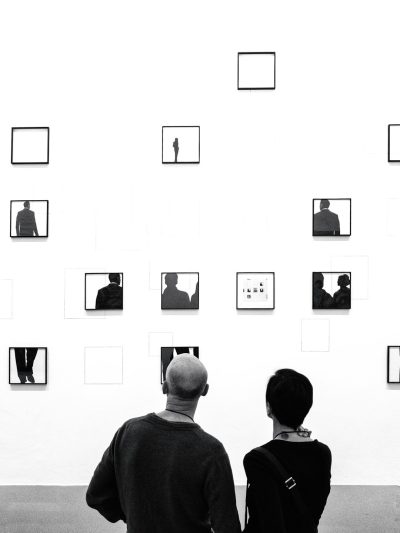 Black and white photograph of two people in an art gallery, looking at the wall with small black frames containing simple black silhouettes on them. The background is white with a minimalist aesthetic. In front there is one man wearing a sweatshirt and a woman with short hair, seen from behind. There should be no shadows or reflections in the style of the photograph. --ar 3:4