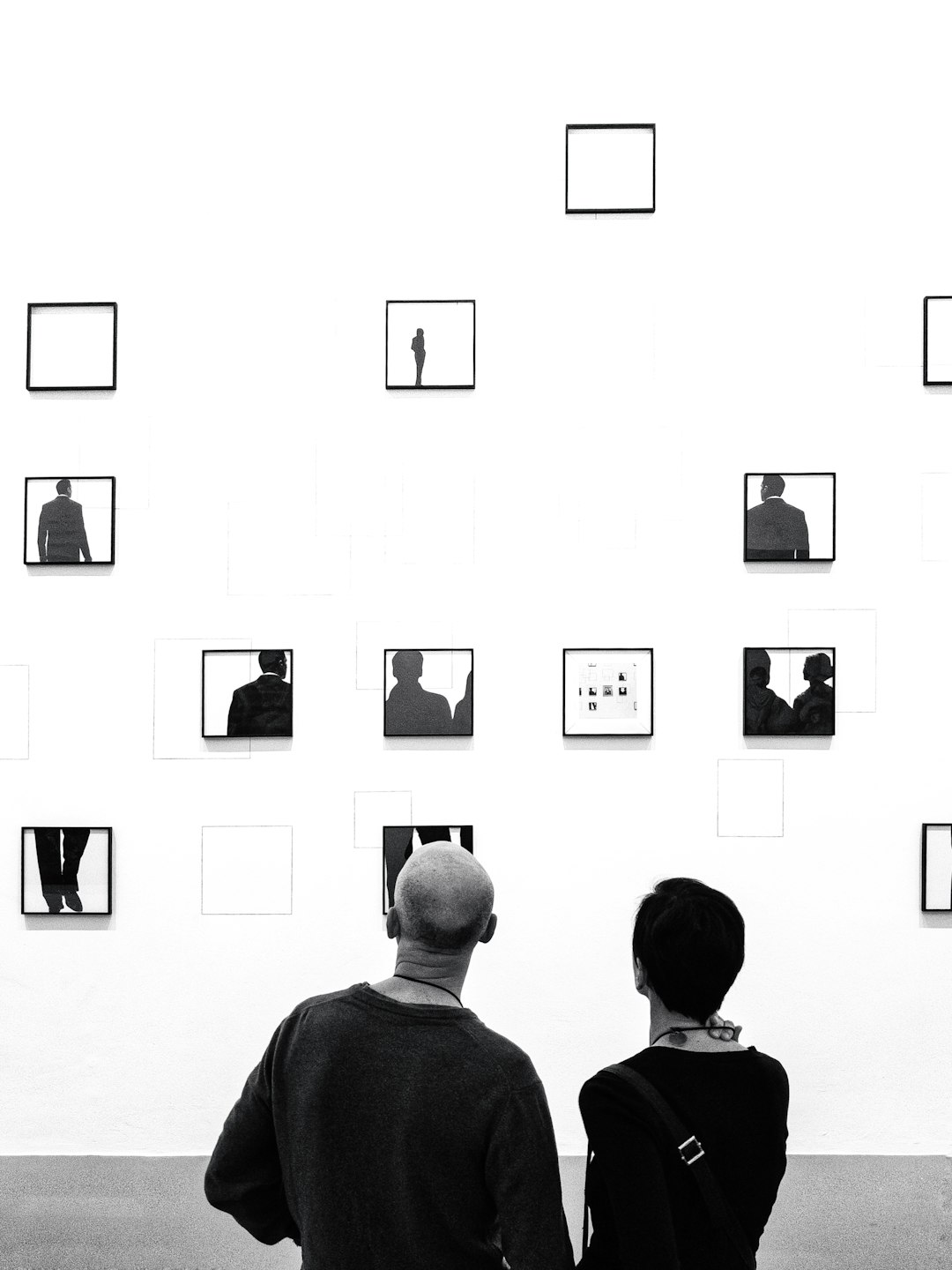 Black and white photograph of two people in an art gallery, looking at the wall with small black frames containing simple black silhouettes on them. The background is white with a minimalist aesthetic. In front there is one man wearing a sweatshirt and a woman with short hair, seen from behind. There should be no shadows or reflections in the style of the photograph. –ar 3:4