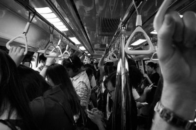 Black and white photography of the interior of an electric train with many people standing and holding onto hand bars. One person is sitting down on his phone looking at it, while another woman stands in front of him wearing earphones and dancing. It is a crowded scene with Asian faces and many legs visible. --ar 128:85