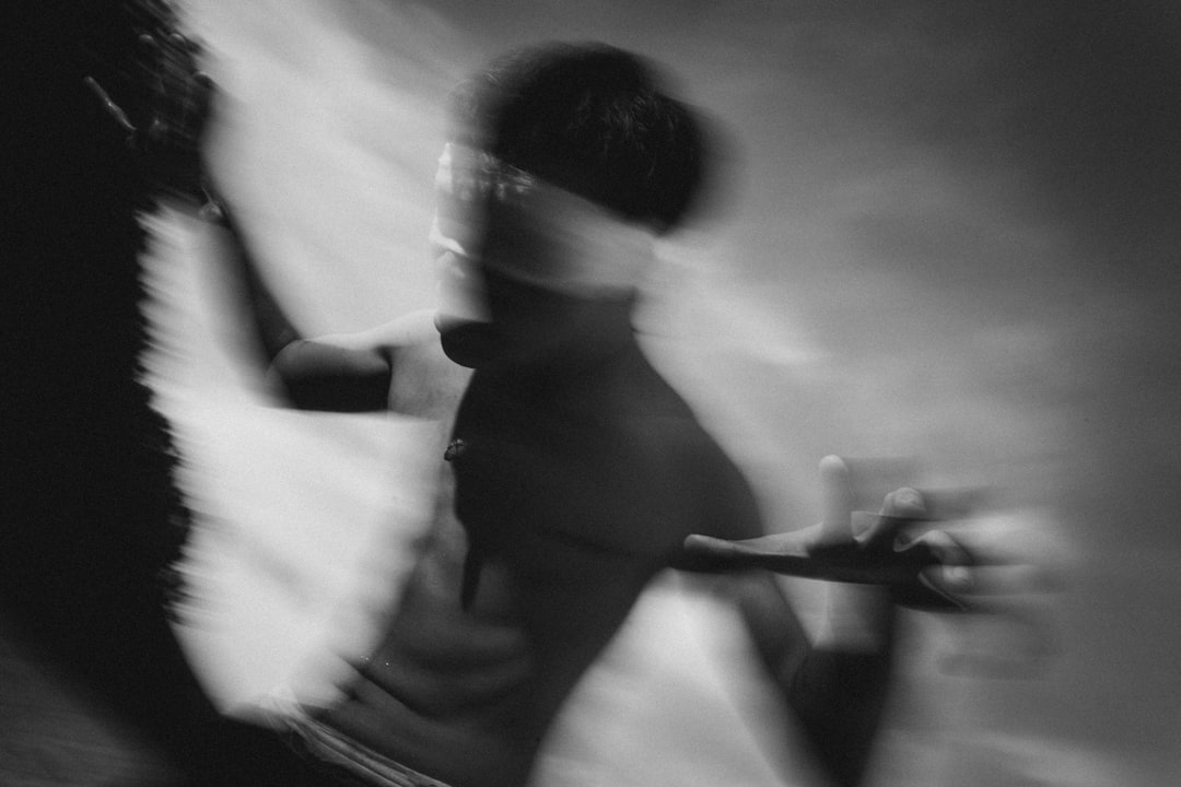 A blurry black and white photo shows two people dancing, with one person’s face obscured by the other person’s hand. The background is blurred to focus on their motion. They appear graceful against an abstract background. Shot from a low angle using a Fujifilm XT4, with an aperture of f/2.0, shutter speed of 1/50, and a 50mm lens set to pan during a slow exposure. A soft light illuminates them, creating a dreamy atmosphere. The photo is in the style of abstract expressionist dance photography. –ar 128:85