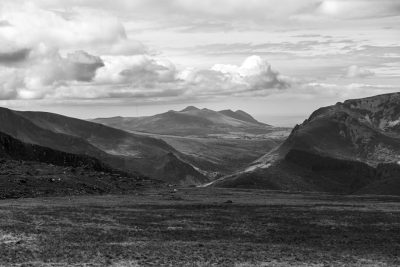 Black and white photography of the mountains in the Scottish volcano landscape with rolling hills, valleys, and distant peaks. In the foreground is an open field leading to rugged mountainous terrain, with clouds scattered across the sky. Shot in the style of Frank sliced. --ar 128:85