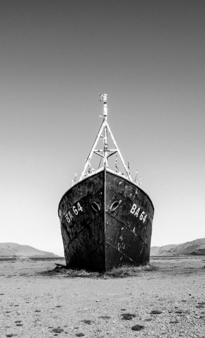 A black and white photograph of an old fishing boat on the ground in the middle of nowhere, with "B64" written on it. The scene is captured from eye level to emphasize its size against the vast landscape around. It's set during daylight with clear skies above, creating a stark contrast between the dark ship body and light background. This composition adds depth by emphasizing details like worn paint, rusted metal, and distant hills. --ar 77:128