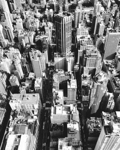 A black and white photograph of the cityscape of New York, with skyscrapers towering over smaller buildings. The photo is taken from an aerial perspective, capturing the bustling streets below. This image symbolizes urban life in big cities, focusing on architectural details like tall structures and narrow alleys. It could be used to represent modern architecture or urban living in the style of an aerial photograph. --ar 51:64