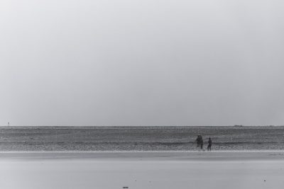 A black and white photograph of two people standing on the beach, fishing in an empty area with flat ground and sand that extends to the far distance, the sky is overcast, the composition should be centered around them, the overall tone must convey tranquility and simplicity, creating a serene atmosphere, shot in the style of a Sony Alpha A7 III camera. --ar 128:85