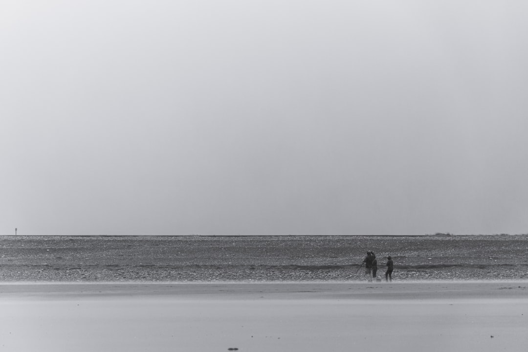 A black and white photograph of two people standing on the beach, fishing in an empty area with flat ground and sand that extends to the far distance, the sky is overcast, the composition should be centered around them, the overall tone must convey tranquility and simplicity, creating a serene atmosphere, shot in the style of a Sony Alpha A7 III camera. –ar 128:85