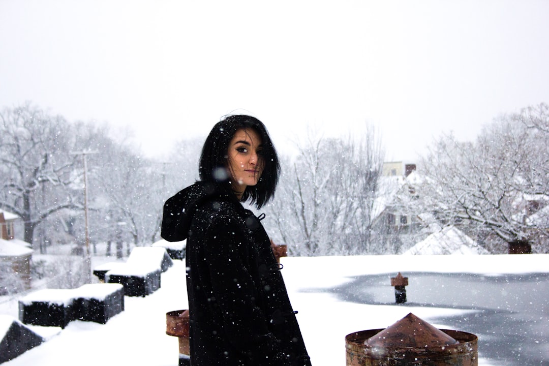 A photo of an attractive Indian woman with black hair standing on the roof in front of her house. It is snowing heavily, with snow falling from above and covering everything. She is wearing all dark  and has short shoulder-length hair. The sky looks grey and the trees behind her have no leaves and look covered in heavy white snow. –ar 128:85