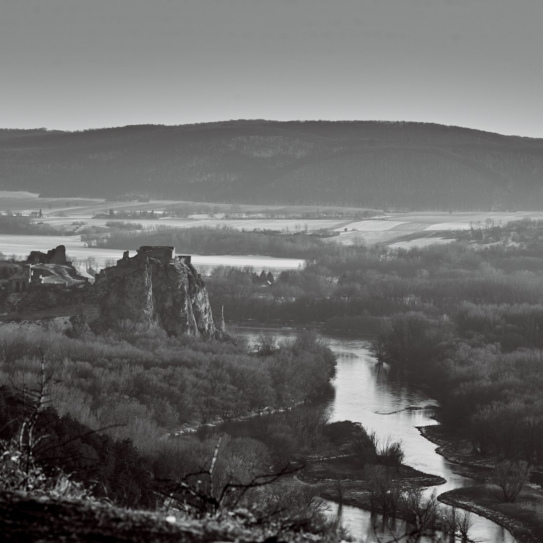 Black and white photograph of the wide river near Chodcontrol in the valley, with rolling hills and an ancient castle visible on top of one hill, taken from high above in the style of Hilla AF Klint. The landscape is serene yet mysterious, bathed in soft morning light.