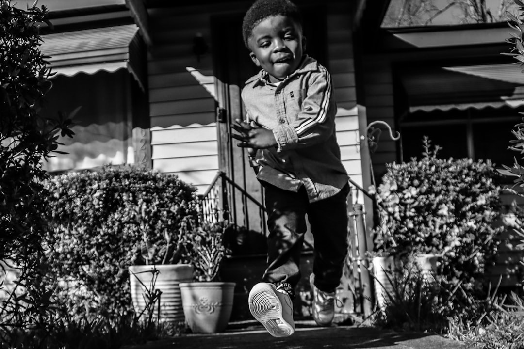 Black and white street photography of a happy black boy wearing sneakers jumping on the front porch, with plants in pots around him, using depth of field, natural light, and a low angle shot. The photo was taken with a Leica Q2 Monochrom and a SummiluxM 50mm f/1.4 ASPH lens. The aperture was set to F8, ISO ranged from 60370 for different lighting conditions, and shutter speed was between. The camera settings can be adapted based on the nostalgic style of a professional photographer. –ar 128:85