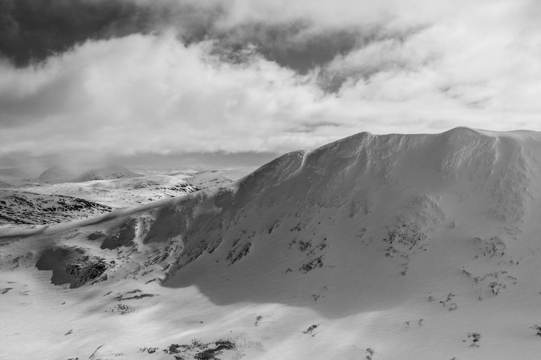 A black and white photograph of the top half of Catfall Mountain in the Cairngorms, with snow covering its peak, taken from an aerial perspective. The mountain is majestic against a cloudy sky, showcasing intricate patterns on its snowy surfaces. A sense of solitude envelops it under soft light. High resolution photography in natural lighting captures global illumination and uplighting, at f/20. –ar 128:85