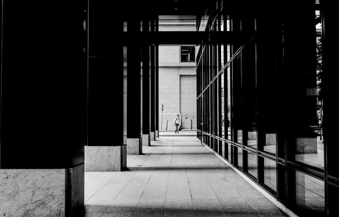 Black and white street photography with high contrast, symmetry, and minimalism. The photo shows a modern architecture with a woman walking in the distance between glass walls in the style of architectural photography. It features a building entrance with reflections on windows and architectural details like shadows of columns in the style of architectural photography. The cityscape background shows an urban environment with contrast lighting and was taken at high resolution with a Canon EOS R5 camera using a macro lens at f/28 aperture and an extremely low ISO value to capture intricate textures and detail. –ar 64:41