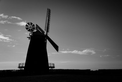 Black and white silhouette of the windmill in Blyth, Northumberland from behind. The sun is shining with some clouds in the sky. Shot on a Nikon D850 camera with an aperture setting of f/2.4, shutter speed of 300mm and a normal ISO level. High resolution, hyperrealistic style. --ar 128:85