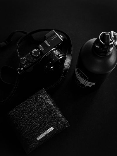 Black and white photography of the product photo, camera with leather strap, black metal bottle in minimalistic modern style, on top is placed an open men's wallet with money inside, all elements lie flat on a dark background, minimalism, minimal design, high resolution, high contrast, high quality, high detail, cinematic light, sharp focus, shot by Hasselblad H6D400c medium format camera --ar 3:4