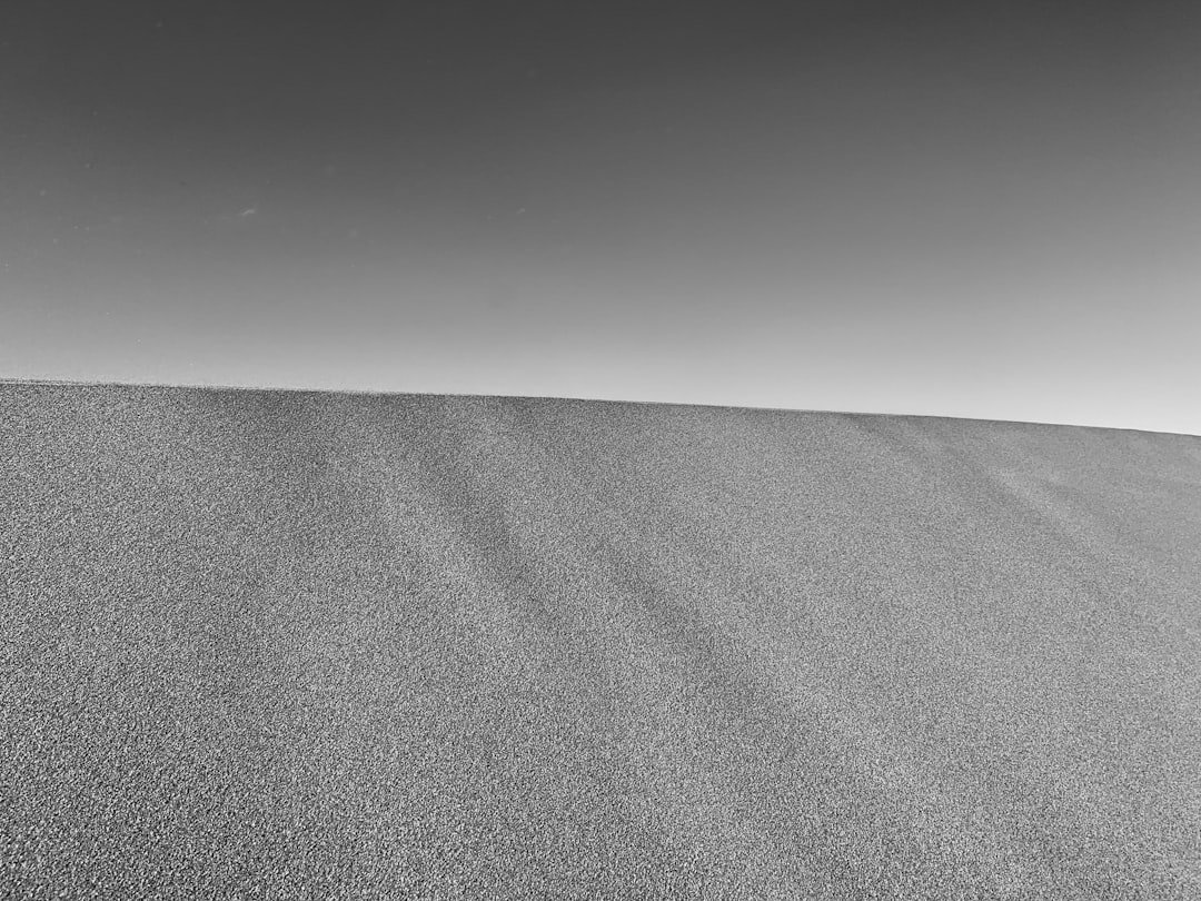 A black and white photograph of the top half of an endless sand dune, with a clear sky in the background. The grainy texture of fine sand is visible against the stark contrast between light gray and dark grey hues. A minimalist composition focusing on simplicity and elegance. –ar 4:3