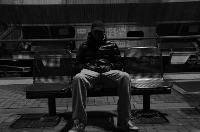 A man sitting on a bench in black and white, street photography, night scene of a city center as he waits for a train, high-definition image quality, photographed in the style of Leica camera with a telephoto lens, symmetrical composition, black cap, gray sweater, jeans pants. The figure is centered with empty space behind it. --ar 32:21