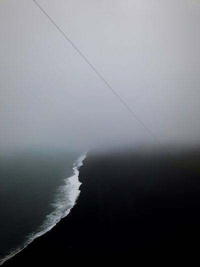 minimalistic aerial photography of the ocean and black sand beach, foggy weather, thin straight line power cable in distance, grey sky, melancholic, sad, melancholy --ar 3:4