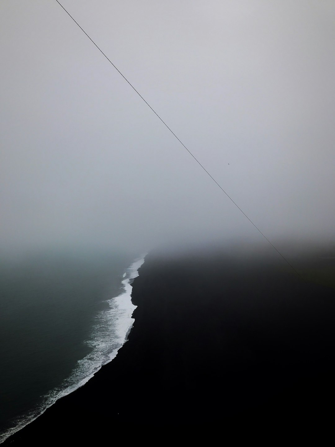 minimalistic aerial photography of the ocean and black sand beach, foggy weather, thin straight line power cable in distance, grey sky, melancholic, sad, melancholy –ar 3:4