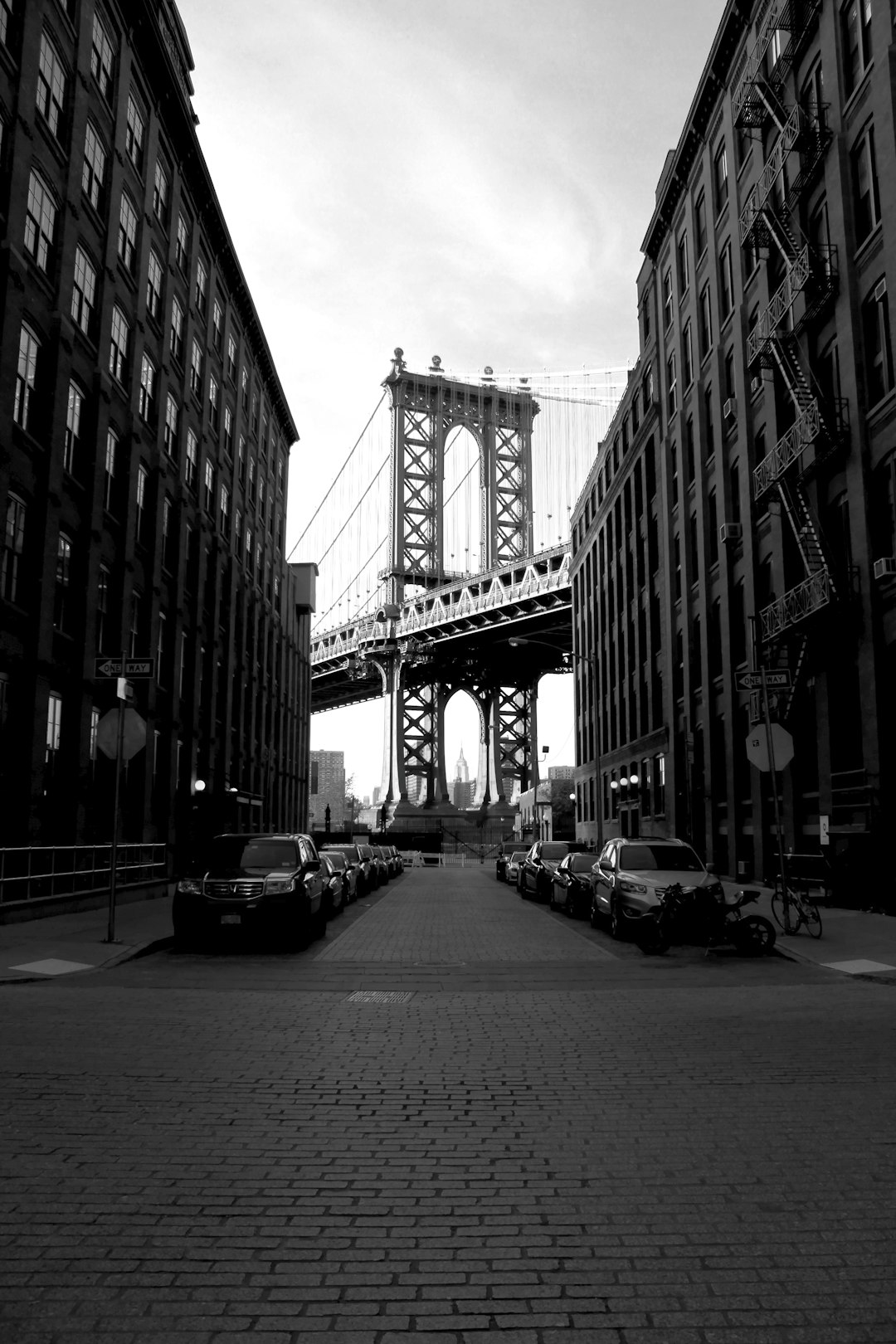 Black and white photography of the Manhattan Bridge in New York City, street view with buildings and cars parked on both sides. The camera is facing towards the bridge. In front there’s an empty cobblestone road leading to the bridge. There should be no people or vehicles in the frame. –ar 85:128