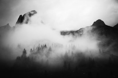 Black and white photography of misty mountains, trees, and clouds in the background. The mountain is covered with dense fog that adds mystery to the scene. There is an open area at its base where part of another forested landscape can be seen. This photo was taken during daylight with a wide-angle lens, giving it a cinematic feel in the style of [Ansel Adams](https://goo.gl/search?artist%20Ansel%20Adams). --ar 128:85