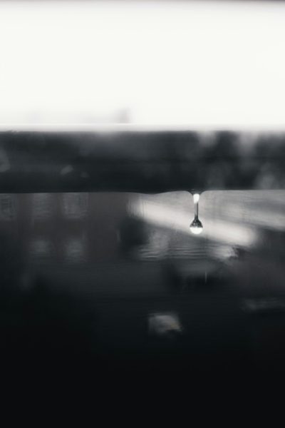 The foreground is blurred, with the edge of an open car window in focus. A single street lamp shines on top of water ripples under it. The overall tone was captured in the style of black and white photography. It has high contrast between lightness and dark tones, presenting soft edges and blurry details. --ar 85:128