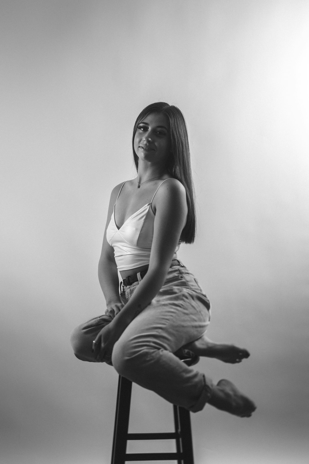 photo of an attractive young adult woman with straight hair, sitting on top and around the edge of a stool in a studio, blackandwhite photography –ar 85:128