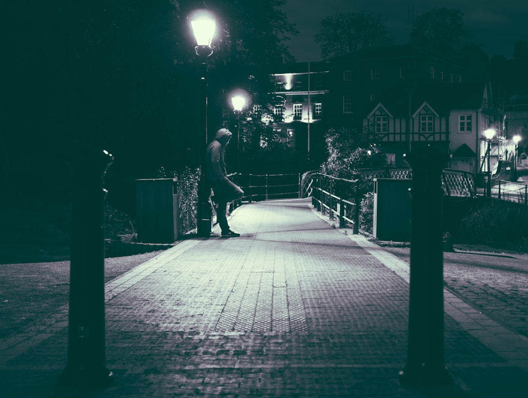 A man walking on the bridge at night, street lights illuminating his path, houses in the background, a dark and mysterious atmosphere, in the style of vintage film camera, wide angle lens, high contrast lighting, a silhouette effect, black and white photography, a night portrait style, in the style of vintage film stock photo. –ar 4:3