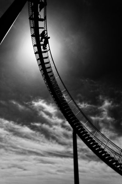 Black and white photography of an extreme roller coaster, with a person climbing the ladder on top and the sun in the background. --ar 85:128