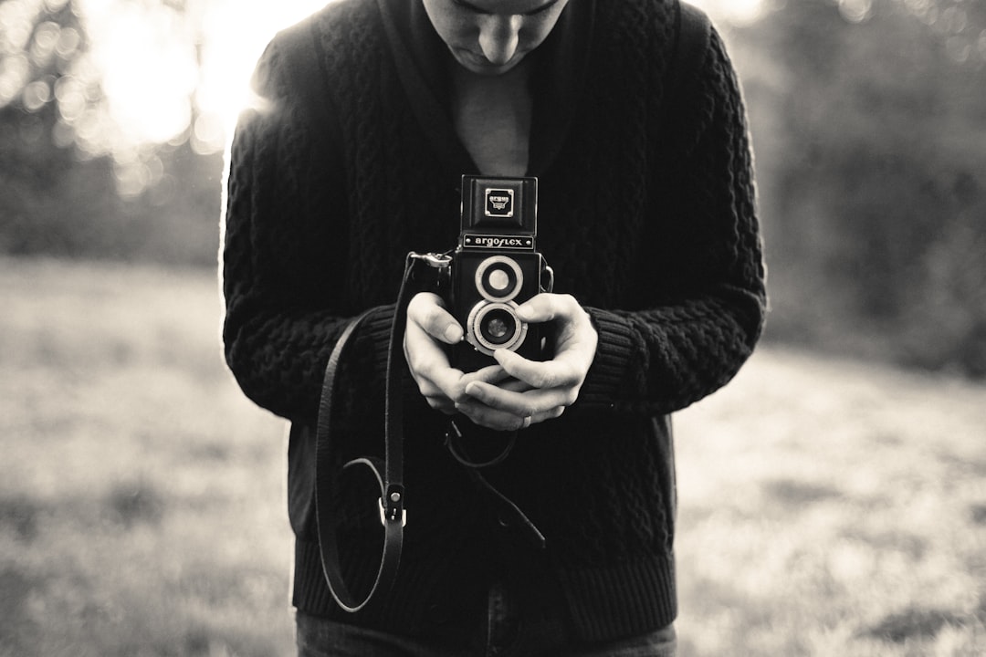 A black and white photograph of an individual holding their vintage film camera, wearing stylish attire with a textured sweater or cardigan. The background is blurred to emphasize the subject, taken during golden hour for warm lighting. Shot from eye level using a Fujifilm GFX 50S, f/2 lens, capturing the intricate details on their hands and , creating a classic and timeless feel in the style of portrait photography. –ar 128:85