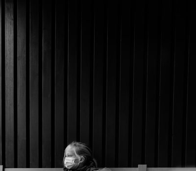 A woman wearing a face mask is sitting on a bench in front of a black vertical slat background, in the style of minimalist photography, in a monochrome style. --ar 128:111