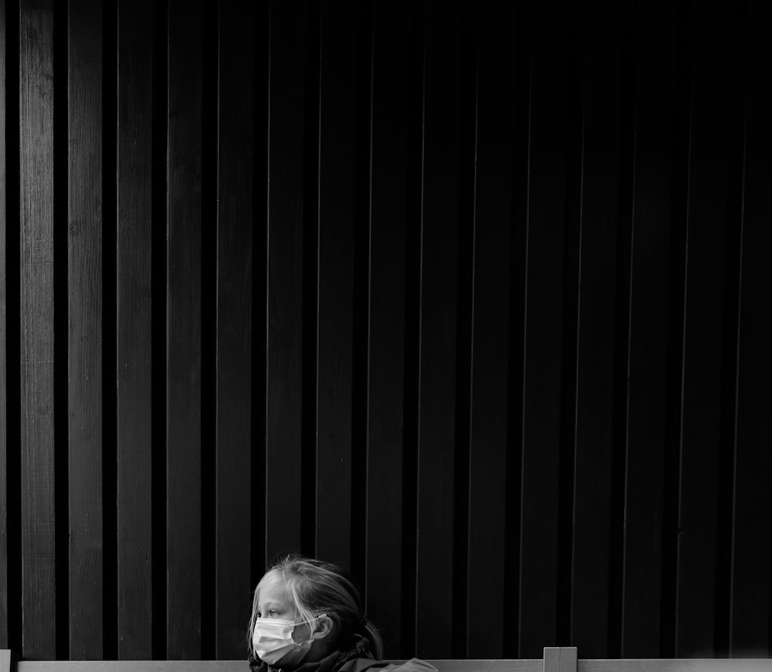 A woman wearing a face mask is sitting on a bench in front of a black vertical slat background, in the style of minimalist photography, in a monochrome style. –ar 128:111