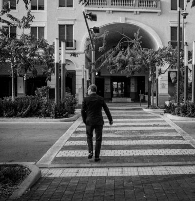 A black and white photo depicts an older man in business attire walking across the street near a mall entrance. He is crossing at the crosswalk in front of a large multilevel commercial building with arched structures and numerous windows. The scene captures him from behind as he walks toward the store entrance, embodying urban life in South Florida at noon. It is shot at eye level, showcasing details like his suit jacket, shoes, and surrounding environment in the style of an urban photographer. --ar 123:128