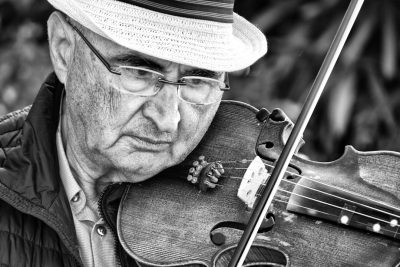 Black and white photo of an elderly man playing the violin, closeup shot in an outdoor setting. Taken with a Nikon D850, focusing on the detailed facial expression and musical instrument details in the style of the man playing. --ar 128:85