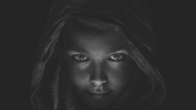 A young girl with dark eyes wearing an open hooded cloak with a nose piercing against a black background in a symmetrical composition with high contrast and a monochrome color scheme with soft lighting in the style of portrait photography creating a mysterious atmosphere. --ar 16:9