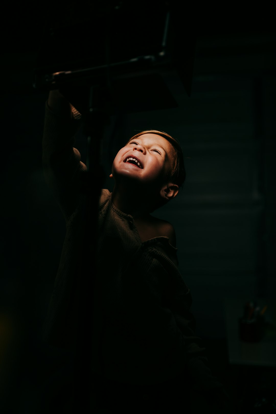 A boy reaching up to the light, smiling in darkness, low angle shot, portrait photography, Canon EOS R5 F2 ISO300, in the style of Canon EOS R5. –ar 85:128